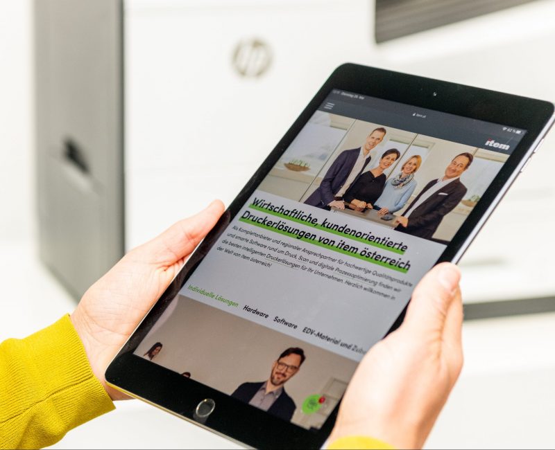 das Bild zeigt ein Tablet mit der item österreich Homepage, im Hintergrund ein HP Drucker