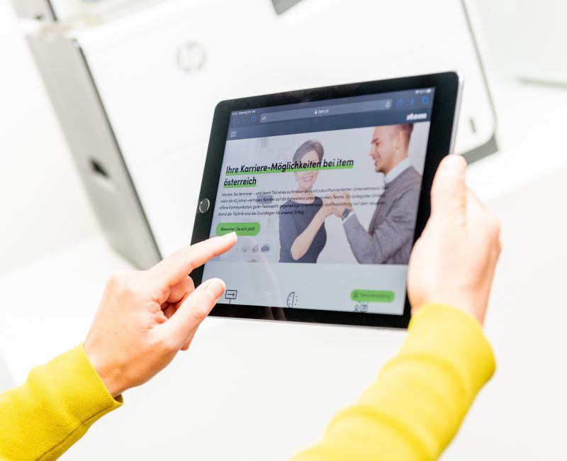 das Bild zeigt ein Tablet, wo die Homepage der item österreich abgebildet ist. im Hintergrund ein HP Drucker