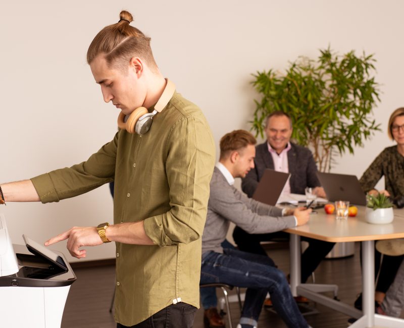 das Bild zeigt einen Mann der einen Drucker bedient. Im Hintergrund, 4 Personen die ein Meeting halten